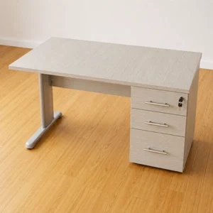 Escritorio en madera clara con cajonera lateral de tres gavetas, cerradura y pata metálica sobre piso de madera.